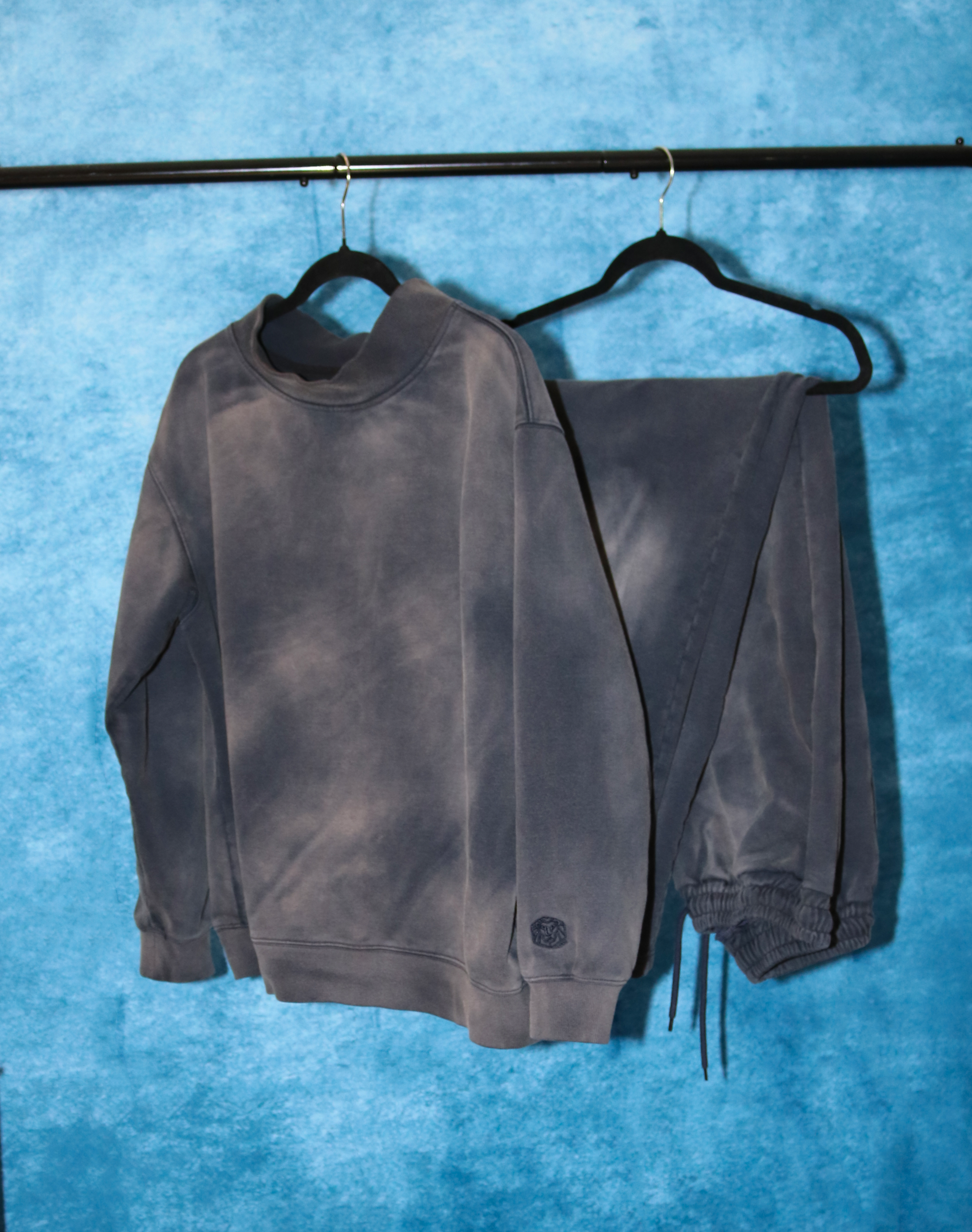 Navy acid wash sweatshirt and pants set on hangers against a blue textured background. Mock Neck Cut. Acid Wash Mock Neck